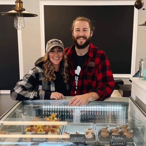 A smiling couple stands behind a dessert display, wearing matching plaid shirts and a cap, with various treats visible.