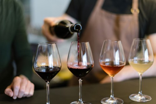 A person pours red wine into a glass, surrounded by three other wine glasses with different types of wine.