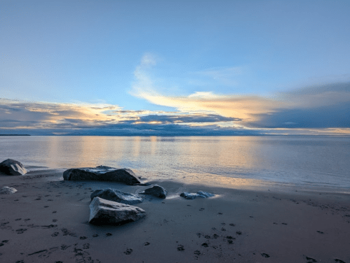 A serene beach scene at sunset, featuring calm waters, scattered rocks, and a colorful sky with clouds.