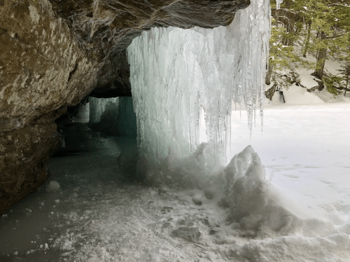 Icicles hang from a rocky cave entrance, with snow and ice covering the ground in a winter landscape.