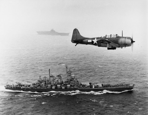 A vintage black and white photo of a battleship sailing below a military aircraft flying overhead.