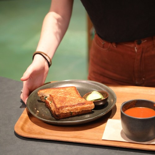 A person serves a plate with grilled cheese sandwiches and a small bowl of tomato soup on a wooden tray.