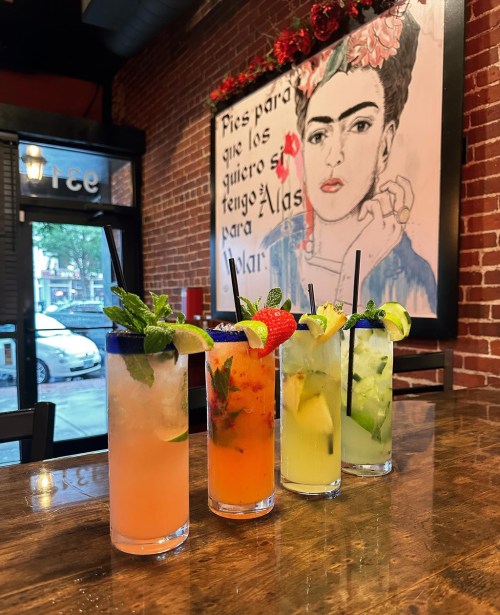 Four colorful cocktails with garnishes sit on a wooden table, against a brick wall featuring a Frida Kahlo mural.