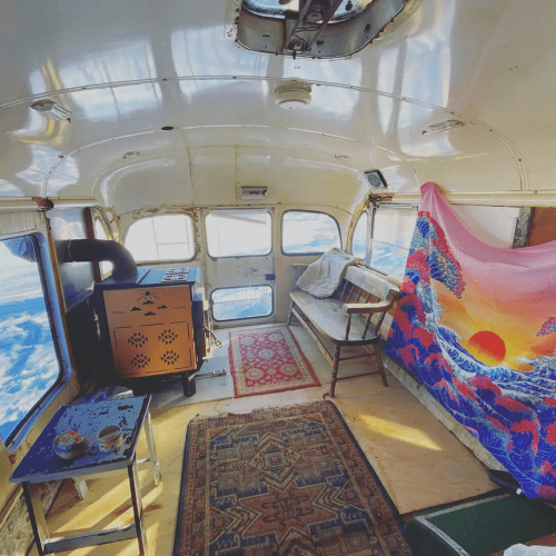 Cozy interior of a converted bus with a wood stove, seating area, and colorful tapestry against a snowy backdrop.