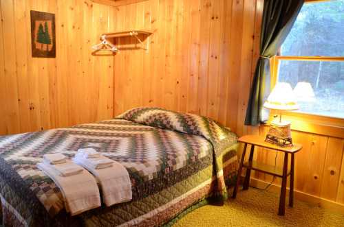 Cozy wooden bedroom with a quilted bedspread, towels, and a lamp beside a window.