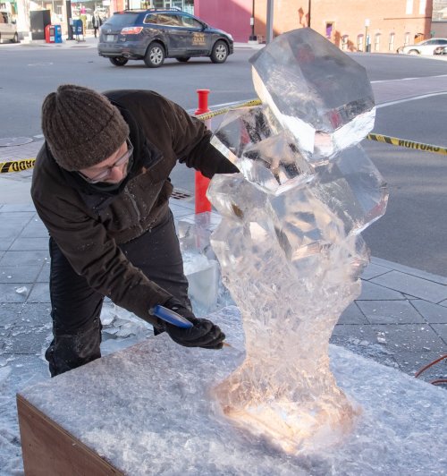 A sculptor chisels an ice block into a creative shape on a city street, surrounded by snow and caution tape.