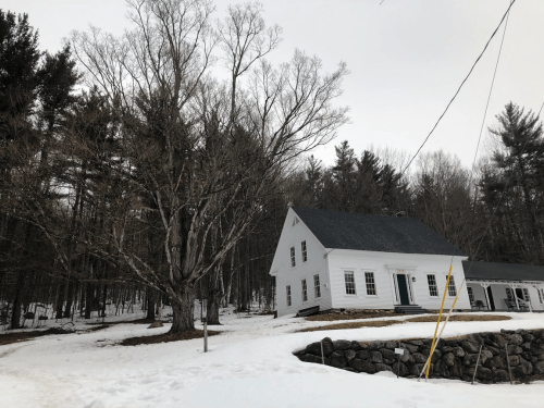 A white house with a dark roof sits near a large tree, surrounded by snow and a forest of bare trees.