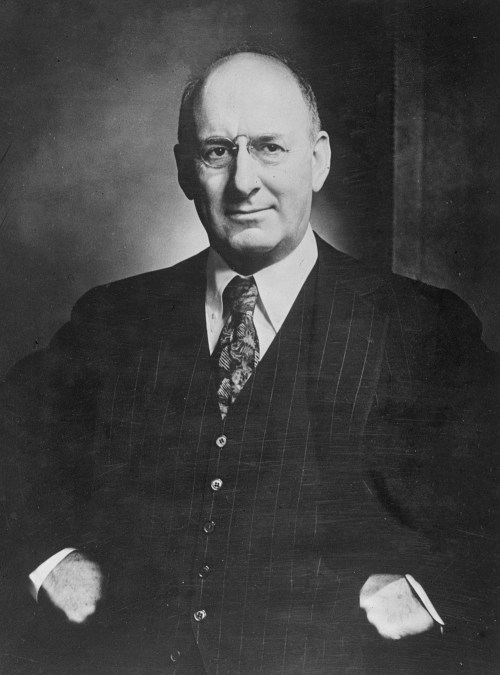 Black and white portrait of a man in a pinstripe suit, wearing glasses, with a serious expression.