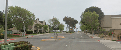 A quiet street lined with trees, leading to a railroad crossing and distant ocean views under a cloudy sky.
