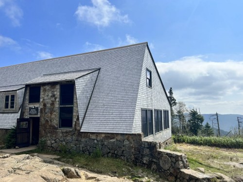 A stone and wood building with a sloped roof, surrounded by trees and mountains under a partly cloudy sky.