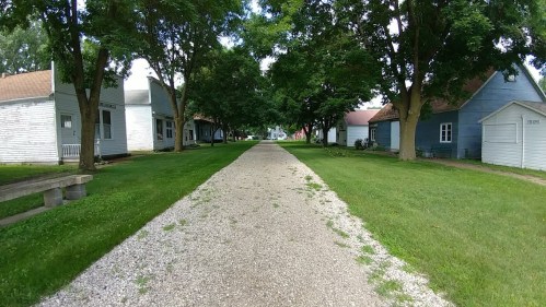 A gravel path lined with trees, leading to buildings on either side in a peaceful, green setting.