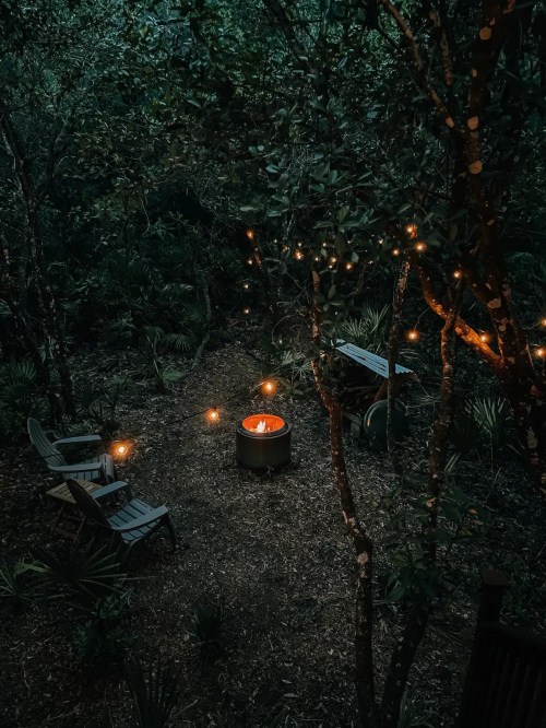A cozy outdoor scene at night with a fire pit surrounded by chairs and string lights in a wooded area.