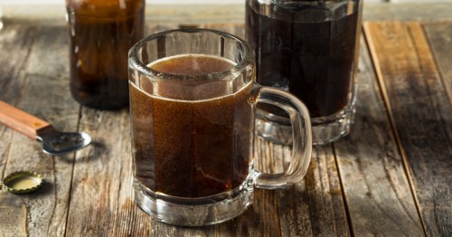 A frosty glass mug filled with dark beer, with a second glass and a bottle in the background on a wooden table.