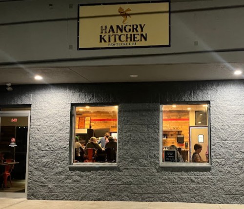 Exterior view of Hangry Kitchen in Pawtucket, RI, with warm lighting and diners visible through the windows.