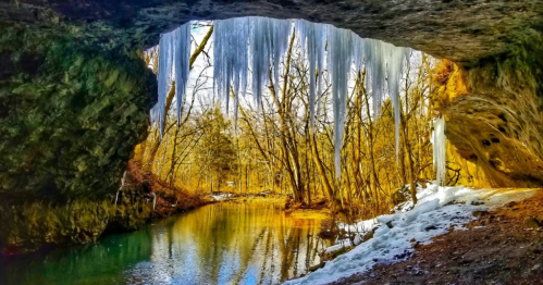 A serene view of a cave opening with icicles, trees, and a calm river reflecting the winter landscape.