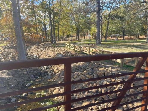 A view of a park with trees, a wooden bridge, and a rocky stream under a clear blue sky.