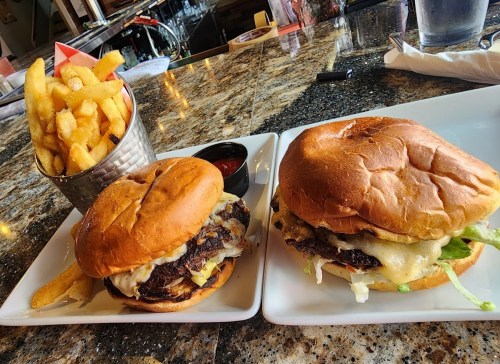 Two burgers with melted cheese and toppings, served with a side of crispy fries in a metal cup.