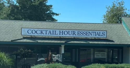 Sign on a building reads "Cocktail Hour Essentials," with greenery in the foreground and a clear blue sky.