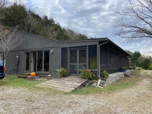 A modern, dark wooden house with a screened porch, surrounded by greenery and a gravel path.