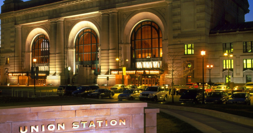 Union Station at night, showcasing its grand architecture and illuminated windows, with parked cars in the foreground.