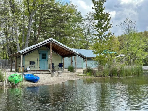 A serene lakeside cabin with a porch, kayaks by the water, surrounded by trees and a cloudy sky.
