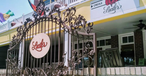 Wrought iron gate with "Lucile's" sign, leading to a colorful restaurant with outdoor seating and flags.