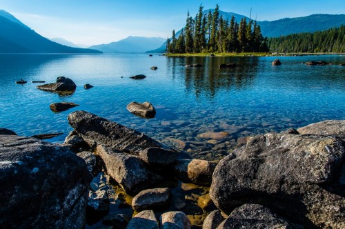 A serene lake scene with clear water, rocky shore, and an island surrounded by trees under a blue sky.