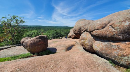 A rocky landscape with large boulders and a clear blue sky, surrounded by green hills in the background.