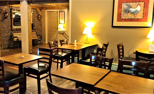 Cozy restaurant interior with wooden tables, chairs, and warm lighting, featuring a decorative rooster painting on the wall.