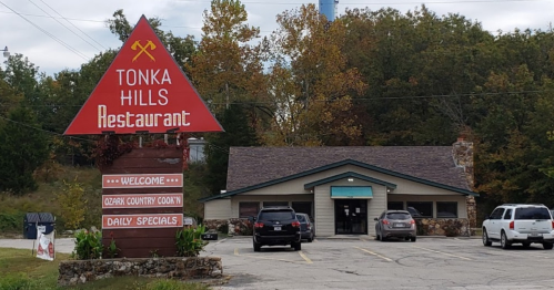 A red triangular sign for Tonka Hills Restaurant with a stone building and parked cars in a wooded area.