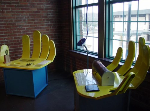 Two colorful interactive exhibits with yellow hands and a cylindrical device, set against a brick wall and large windows.