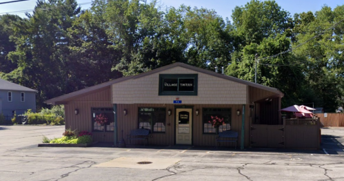 A cozy tavern building with a sign, surrounded by trees and a parking lot, featuring outdoor seating.