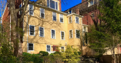 A yellow multi-story house surrounded by trees and greenery, with large windows and a red door.