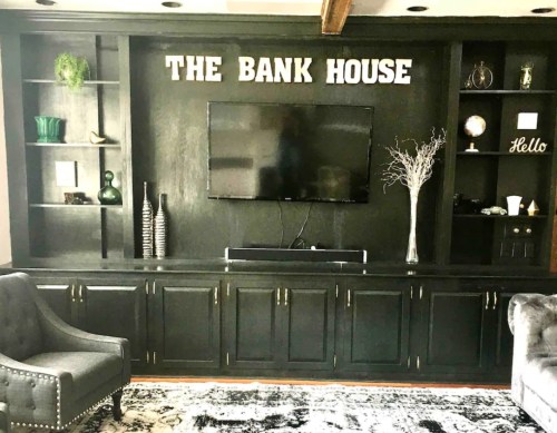A stylish living room with a black entertainment unit, TV, decorative shelves, and the words "THE BANK HOUSE" on the wall.