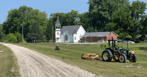 A rural scene featuring a church, a tractor, and a lawn mower on a grassy area beside a gravel road.