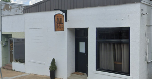 Exterior view of a building with a sign reading "Jail Hotel," featuring a simple white facade and a small plant by the entrance.