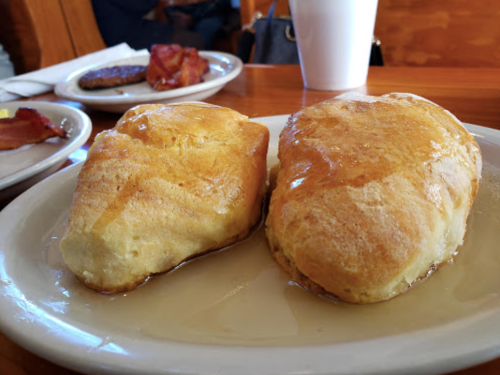 Two golden, fluffy biscuits drizzled with syrup on a white plate, with a blurred background of breakfast items.