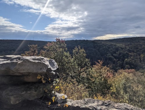 A rocky ledge overlooks a forested valley under a partly cloudy sky, with autumn foliage in vibrant colors.