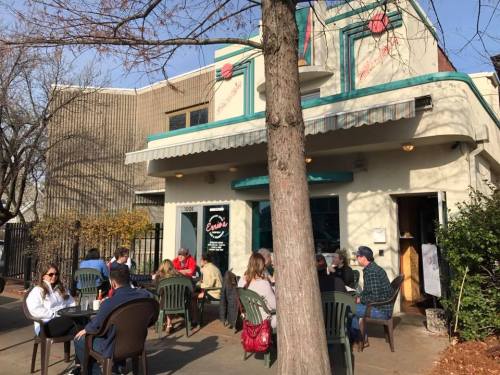 A cozy café with outdoor seating, patrons enjoying drinks, and a tree in the foreground on a sunny day.