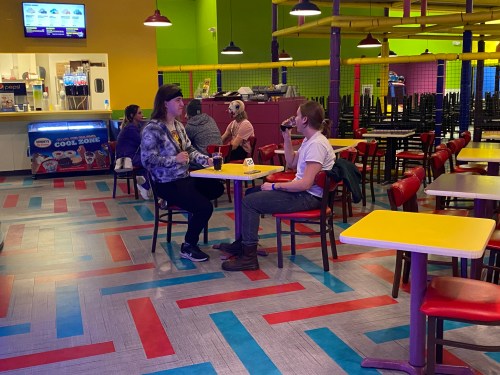 Two people sit at a colorful table in a vibrant indoor space, chatting and enjoying drinks. Bright decor surrounds them.