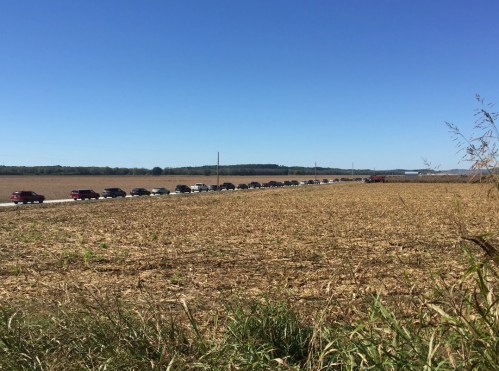 A long line of cars on a road beside a harvested cornfield under a clear blue sky.