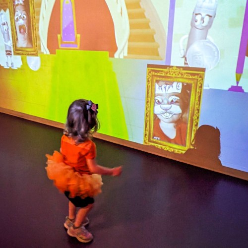 A child in an orange tutu interacts with colorful animated projections on a wall, featuring playful characters.