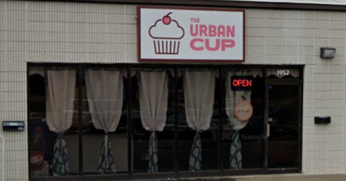 Exterior of "The Urban Cup" bakery with a cupcake sign and open neon light, featuring large windows and curtains.