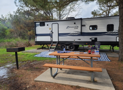 A camping setup featuring a Zinger travel trailer, picnic table, and grill in a wooded area.