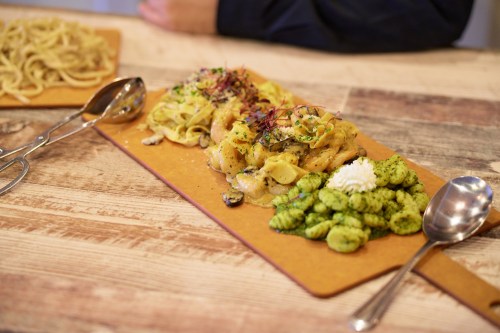 A wooden table with two plates of pasta: one with creamy fettuccine and the other with green gnocchi and herbs.