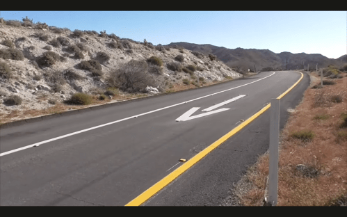 A winding road with a yellow arrow marking, surrounded by rocky terrain and sparse vegetation under a clear blue sky.