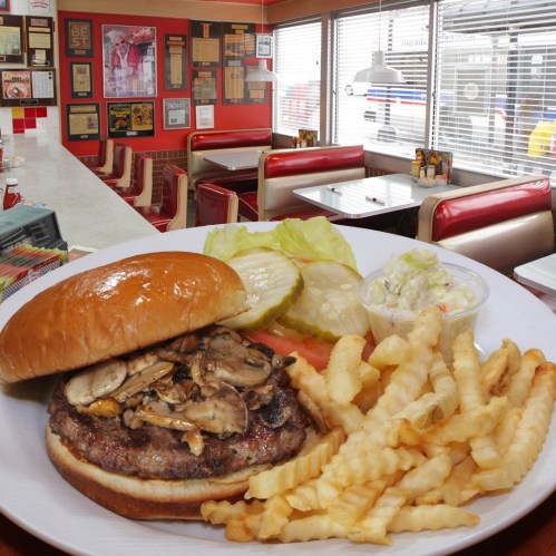 A juicy burger topped with mushrooms, served with lettuce, pickles, coleslaw, and crispy fries in a retro diner setting.