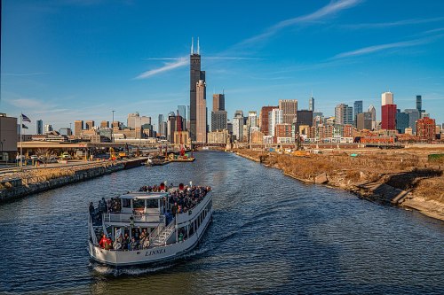 A boat with passengers navigates a river in Chicago, with the skyline and tall buildings in the background under a clear blue sky.