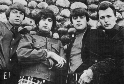 Four men in 1960s fashion pose against a stone wall, exuding a cool, confident vibe.