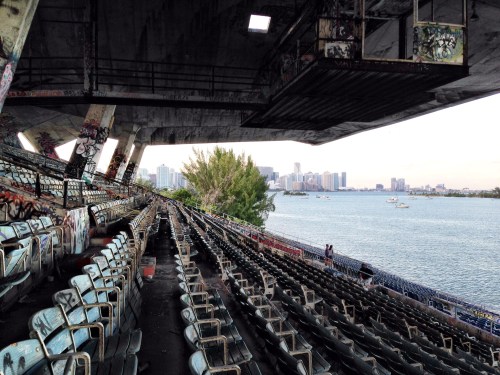 Abandoned stadium seating with graffiti, overlooking a waterfront city skyline and trees in the background.